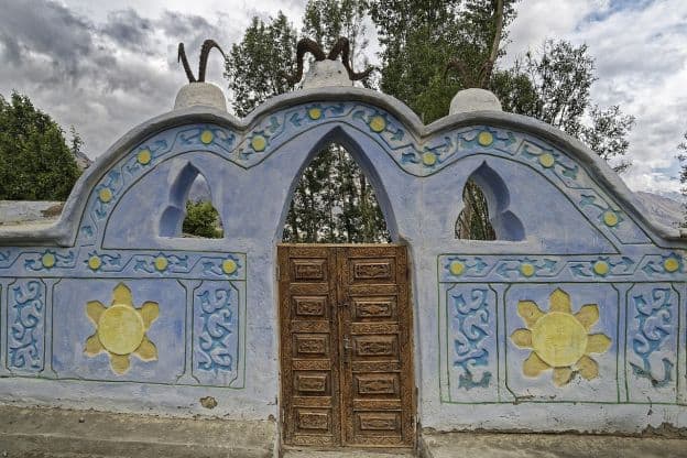 A decorated blue stone wall in Vrang, Tajikistan. There is a set of wooden doors in the middle of the wall, surrounded by arches and carvings that look like flowers or suns. Three sets of antlers or horns top the wall.