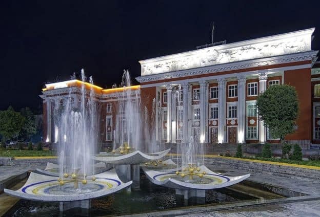 Three water fountains run at night in front of the Houses of Parliament in Dushanbe, Tajikistan's capital. The Parliament building, with neoclassical architecture, is painted a reddish color.