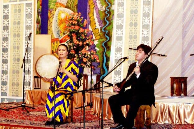 A female and a male folk performer sit next to each other on a colorful stage. The woman is holding what appears to be a drum, and the man has a stringed instrument.