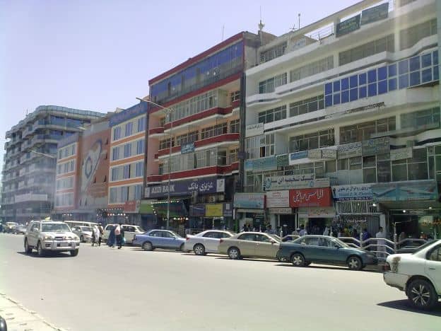Part of a city block in a commercial district in Kabul, Afghanistan. There are shops in large buildings, with several cars parked out front and shoppers walking down the street.