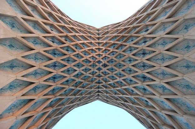 A view of the Azadi Tower, a famous Tehran landmark, from beneath the center arch. The crisscrossing of vaulted compartments, decorated with intricate blue patterns, is visible.