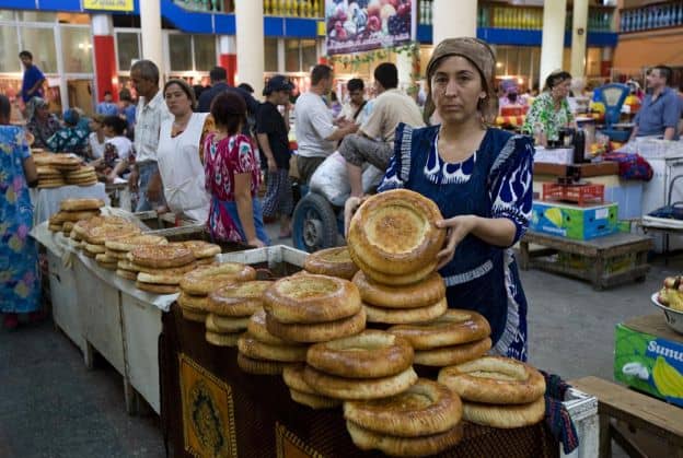 A Tajik woman holds up a round loaf of bread. She is standing in front of a table filled with multiple loaves of similar bread at a marketplace. There are many other people in the background.