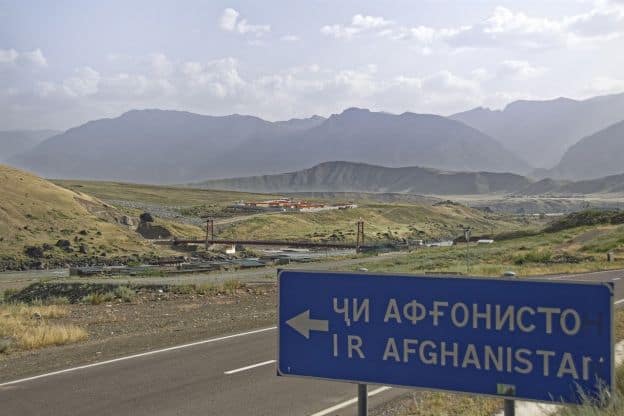 Road sign in Tajiki, with secondary text in the Latin alphabet, indicating the border between Tajikistan and Afghanistan. The Friendship Bridge over the Panj River is shown in the background.