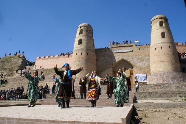 A group of folk performers in traditional Tajik costumes performs outdoors at Hissar Fortress, an ancient building with high walls and towers. There appear to be both dancers and musicians in the group.