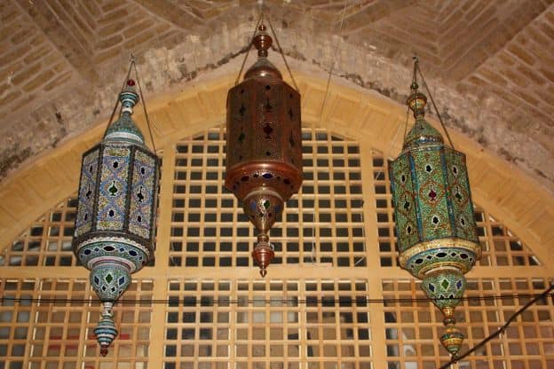 Three ornate lamps hang from a brick ceiling in front of a wooden screen at a mosque in Isfahan, Iran.