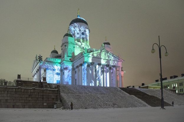 Against a stark background of a tawny-gray sky, the white façade of Helsinki Cathedral is vividly colored with pictures by a projected light show. The Cathedral looms above a steep double-flight of snowy stairs.