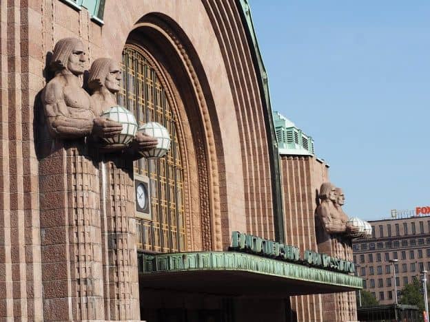 The art-deco Helsinki Central Railway Station is made of reddish-brown stone. Four large statues of men bearing globes of light flank the arched central entryway, from which projects an oxidized metal awning.