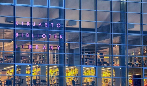 A view of the glass-covered walls of a city library in Oulu, Finland. Multiple stories of the library stacks are visible, as are some patrons. The library is identified by a purple neon sign in Finnish.