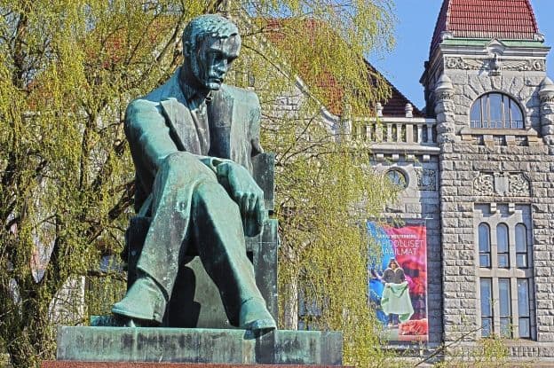 An oxidized bronze statue of author Aleksis Kivi in a contemplative pose. There is a tree and a stone railway building behind the statue.