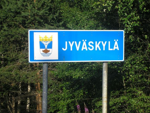 A blue city limits sign for Jyväskylä, Finland stands in front of a group of evergreen trees. The sign also displays Jyväskylä's coat of arms, which features water, a ship, and a Caduceus, topped with a crown.