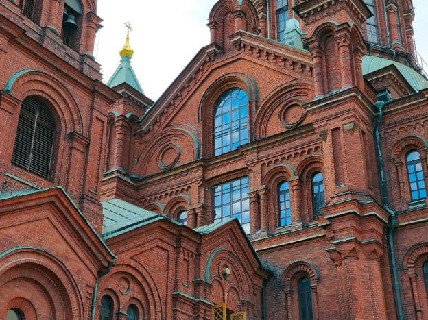 The imposing façade of Uspenski Cathedral is covered in red brick a with light green roof. The architecture is a Romanesque style, with rounded arches surrounding windows and doors.