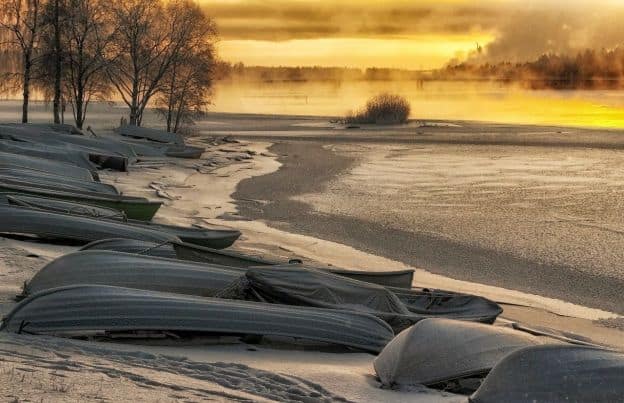 Inverted rowboats line the shore as the sun sets on a beach in Finland. There is snow on the sand.