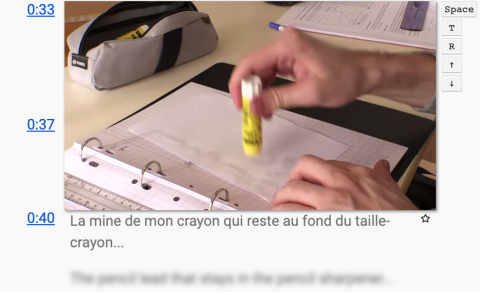 A hand with a glue stick glueing paper in a binder. Time stamps to the right of the image with French subtitles below. English words blurred out below the subtitles. 
